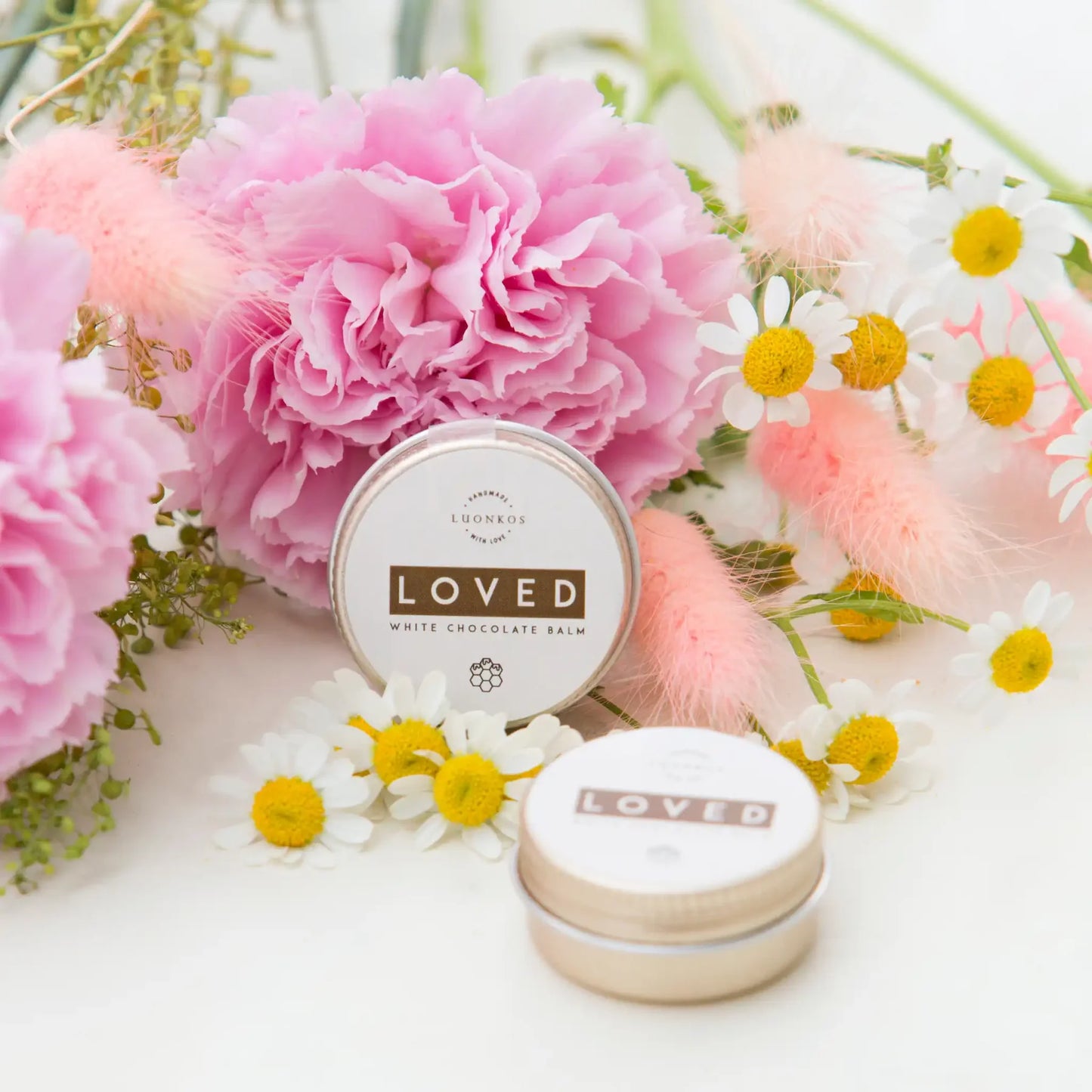 Eine Dose Luonkos Loved White Chocolate Mehrzweck-Lippenbalsam steht zwischen rosa Nelken, Gänseblümchen und zartrosa Blättern, davor eine weitere Dose. Das Arrangement wirkt hell, frisch und blumig.
