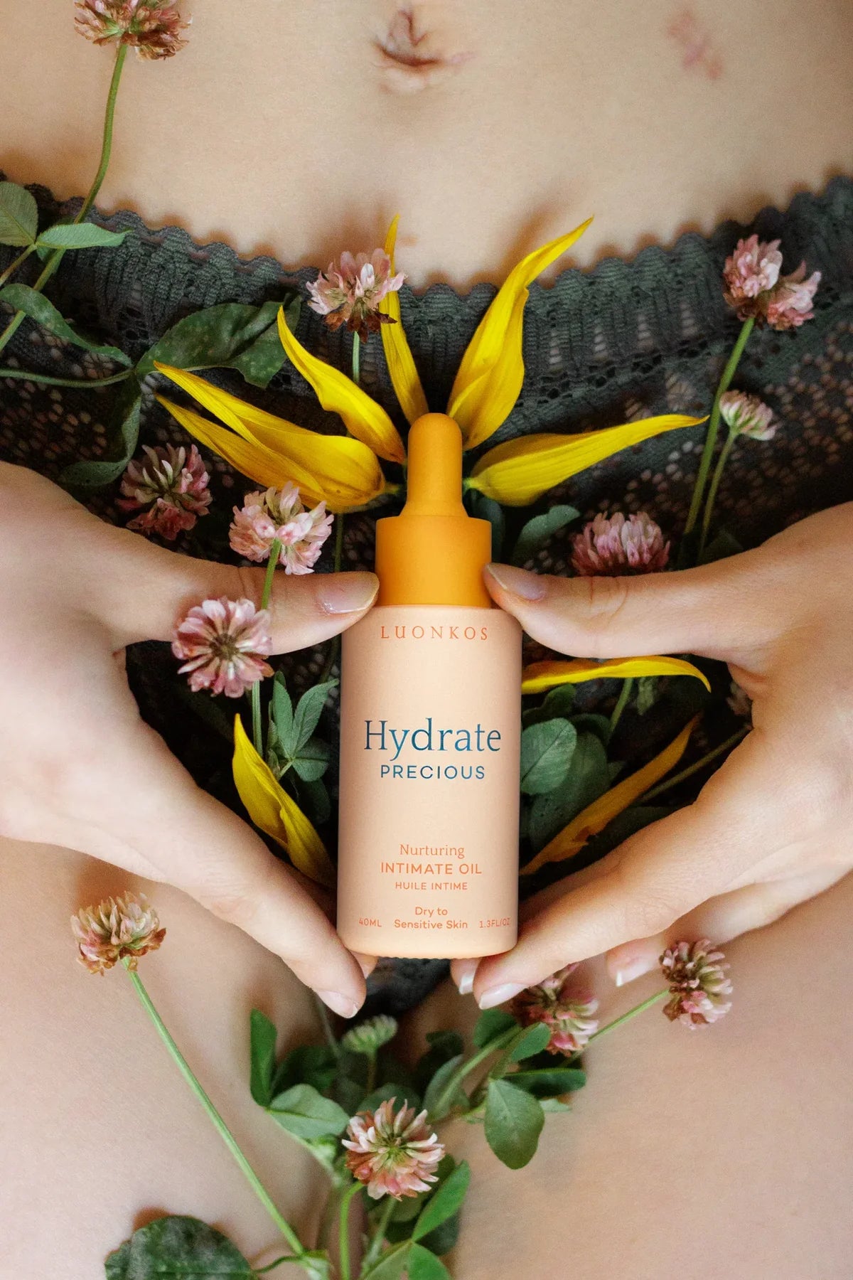 Eine Person hält eine Flasche Luonkos Precious Intimate Oil by Luonkos an ihren Unterleib, umgeben von gelben Blütenblättern und rosafarbenen Kleeblättern, wobei grüne Spitzenunterwäsche sichtbar ist - ideal für empfindliche Schleimhäute.
