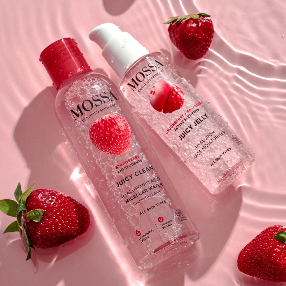 Zwei durchsichtige Mossa-Flaschen aus dem Juicy Berry Gift Set mit feuchtigkeitsspendendem Mizellenwasser und Erdbeer-Illustrationen ruhen auf einer rosafarbenen Oberfläche mit Wasserrippeln. Frische Erdbeeren in der Nähe sorgen für ein fröhliches und sommerliches Flair.