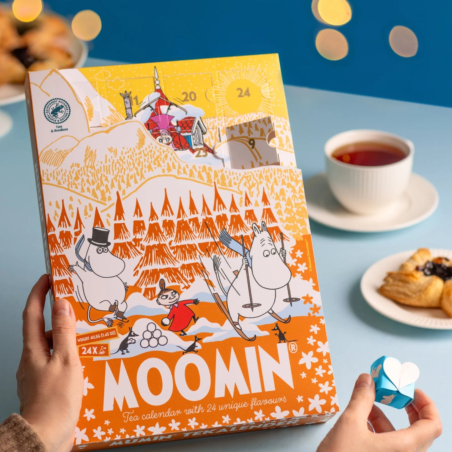Eine Person hält den Nordqvist Mumin-Adventskalender, eine bunte vegane Teeauswahl von Nordqvist, auf einem blauen Tisch mit einer Tasse Tee, Gebäck und einem blauen herzförmigen Origami. Warme gelbe Lichter leuchten im Hintergrund.