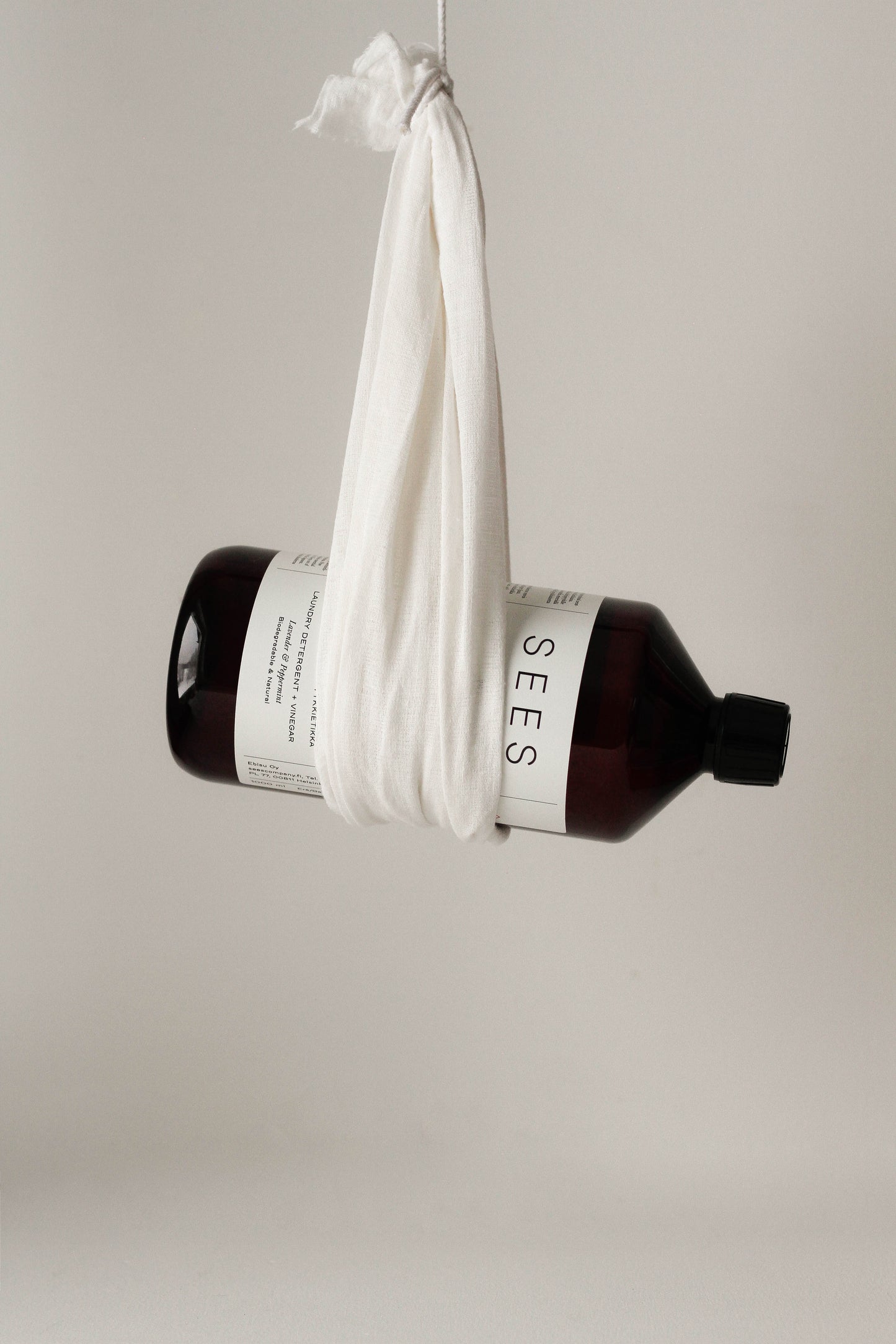 Eine dunkelbraune Flasche SEES Company mit der Aufschrift "SEES 2-in-1 Laundry Detergent with Vinegar" ist in ein weißes Tuch eingewickelt und hängt vor einem schlichten, hellen Hintergrund.