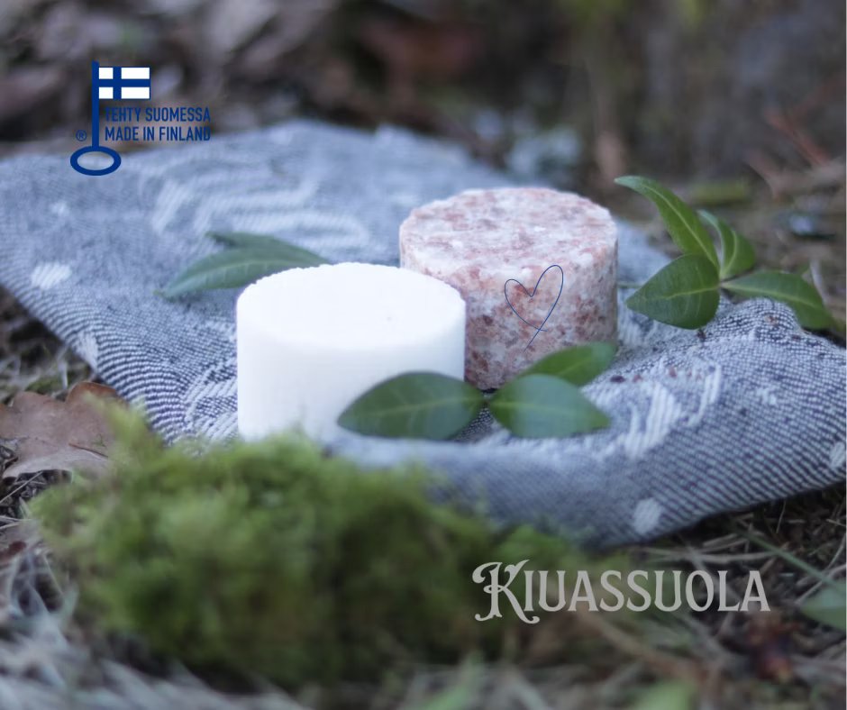 Zwei runde Kiuassuola-Sauna-Salzseifen, eine weiße und eine rosa gesprenkelte, ruhen auf einem gemusterten Tuch im Freien inmitten von grünen Blättern und Moos. Ein "Made in Finland"-Etikett unterstreicht ihre Herkunft und ihre Verbindung zur traditionellen finnischen Saunapflege.
