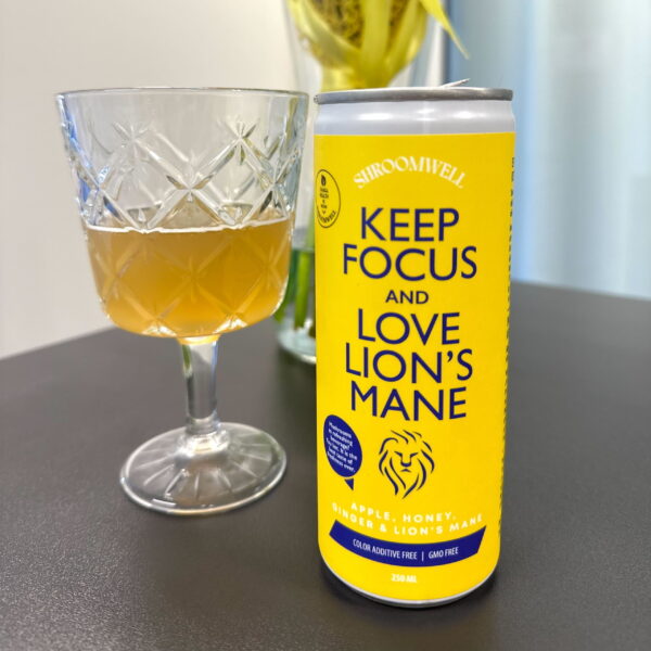 Ein Glas mit einem hellbraunen Getränk steht neben einer gelben Dose des Shroomwell Lions Mane Focus Energy Drink von Shroomwell auf einem dunklen Tisch, auf dem Blumen und ein heller Hintergrund zu sehen sind.