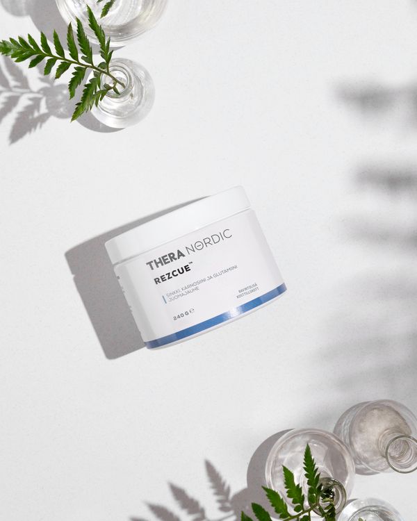 Ein weißer Behälter mit der Aufschrift THERA Nordic Rezcue Drink Powder von THERA Nordic, der für die Darmheilung entwickelt wurde, steht auf einer weißen Fläche, umgeben von Glasvasen mit grünen Farnblättern. Das einfallende Sonnenlicht wirft weiche Schatten.