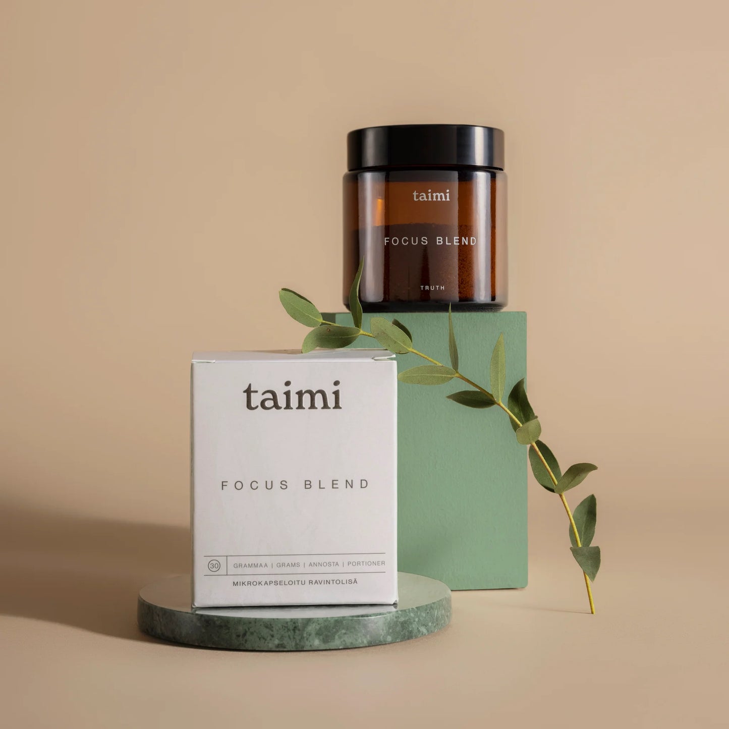 Ein Glas und eine Schachtel mit Taimi Focus Blend von Taimi, einem Nahrungsergänzungsmittel zur Unterstützung der kognitiven Fähigkeiten, werden auf grünen Ständern mit Grünpflanzen vor einem beigen Hintergrund präsentiert. Die minimalistische Verpackung hebt die gehirnfördernden Vorteile hervor.