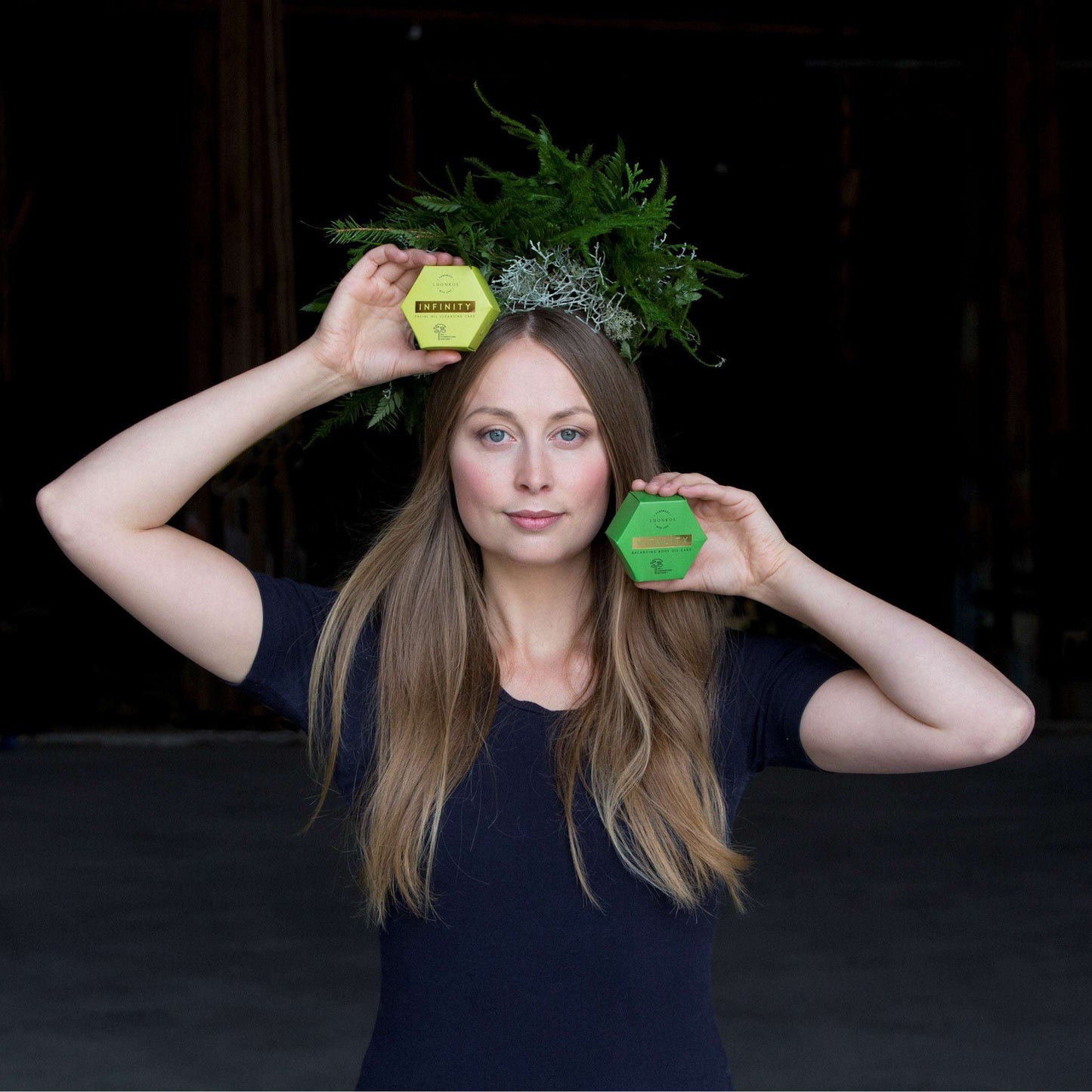 Eine Frau mit langen braunen Haaren und einer Farnkrone posiert vor einem dunklen Hintergrund und hält Sechsecke mit Luonkos Vitality Body Oil Cake und Forest Microbe Extract, um das Mikrobiom der Haut zu feiern.