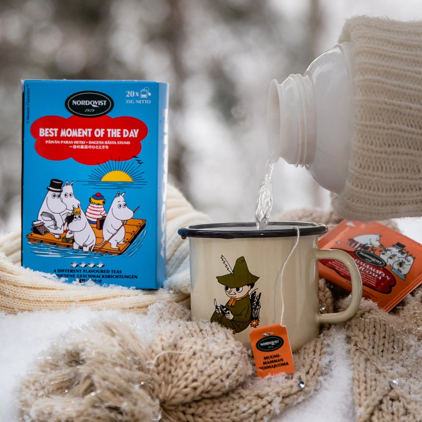 Eine Schachtel Nordqvist Best Moment of the Day Tee liegt auf Schnee neben einer weißen Tasse mit einer Cartoon-Figur. Heißes Wasser tränkt den schwarzen Teebeutel, während ein Schal und Handschuhe an den Genuss gemütlicher Nordqvist-Fruchtmischungen im Freien erinnern.