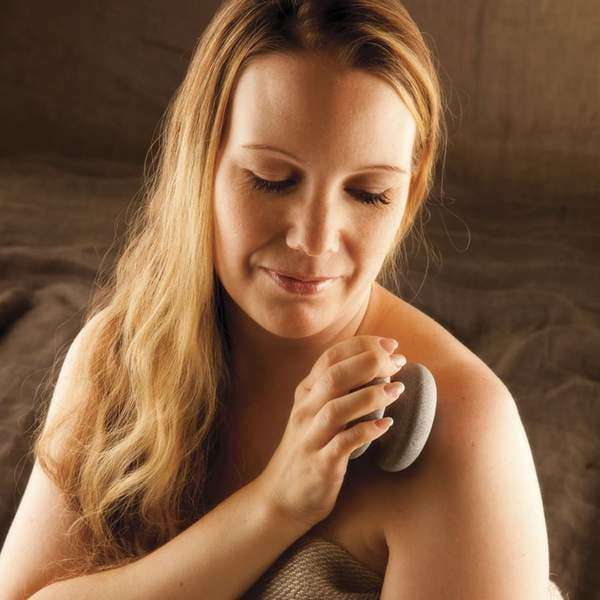 Eine Frau mit langen blonden Haaren peelt sanft ihre Schulter mit dem Hukka Sauna-Joy Massagegerät von Hukka. Sie lächelt sanft und ist in ein Handtuch gehüllt, vor einem gemütlichen, neutralen Hintergrund.