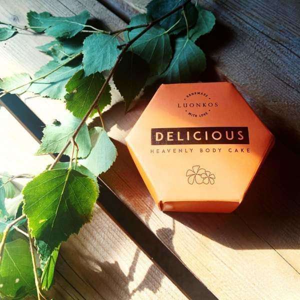 Eine sechseckige orangefarbene Schachtel mit der Aufschrift Luonkos Delicious Body Cake ruht auf Holz neben grünen Blättern im Sonnenlicht und erinnert an die nährende Pflege und die ätherischen Öle dieser natürlichen Körperfeuchtigkeitspflege von Luonkos.