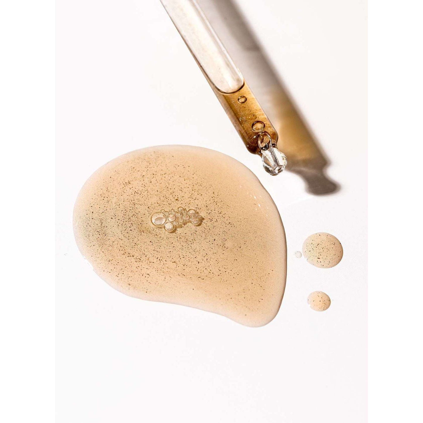 Eine Pipette gibt das Moi Forest Dust Microbiome Magic Oil auf eine weiße Oberfläche ab. Dabei entstehen ein großer und zwei kleine, golden schimmernde Tröpfchen dieses Mehrzweck-Hautöls von Moi Forest, die jeweils mit winzigen Partikeln gesprenkelt sind.