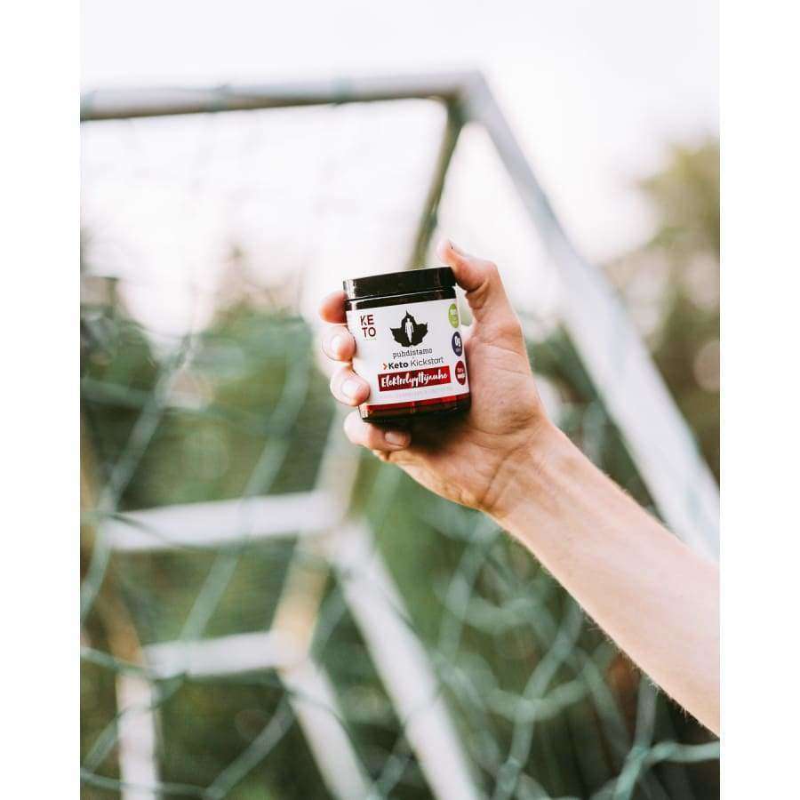 Eine Hand hält ein kleines Glas mit der Aufschrift „Puhdistamo Electrolyte Powder Forest Fruit“ von Puhdistamo vor einem Fußballtor im Freien, was auf die Unterstützung der Flüssigkeitszufuhr hinweist; der Hintergrund ist leicht verschwommen.