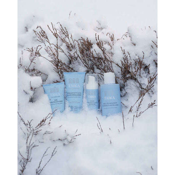 Quatre produits Taika bleu clair, dont le lait démaquillant Taika Sensitive Cleansing Milk, se dressent dans un décor enneigé, entourés de brindilles recouvertes de neige. Idéal pour les peaux sèches ou sensibles, cet arrière-plan hivernal met en valeur ces produits de soin indispensables.