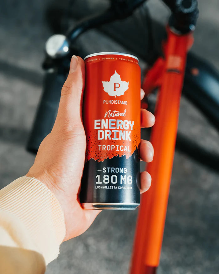 Una mano regge una lattina di Puhdistamo Natural Energy Drink Tropical Strong, con l'etichetta "Strong 180 mg", con una bicicletta arancione sfocata sullo sfondo. Questa bevanda energetica Puhdistamo senza zucchero contiene caffeina proveniente da estratti vegetali.