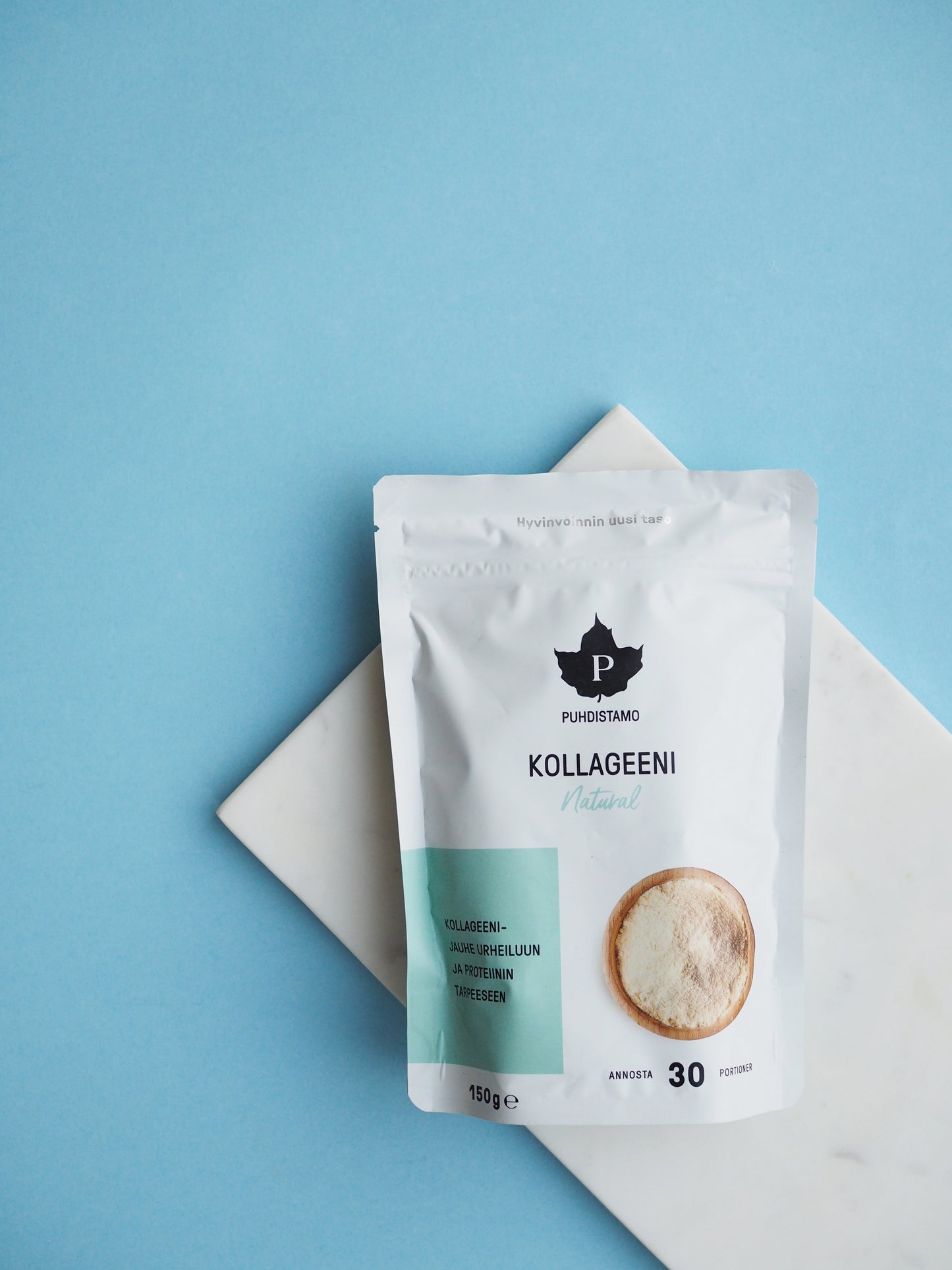 Una bustina bianca di Puhdistamo Collagen Natural di Puhdistamo è appoggiata su una lastra di marmo bianco con sfondo azzurro. La confezione riporta un logo con una foglia nera, testo in finlandese e sottolinea il contenuto di peptidi di collagene Bodybalance.