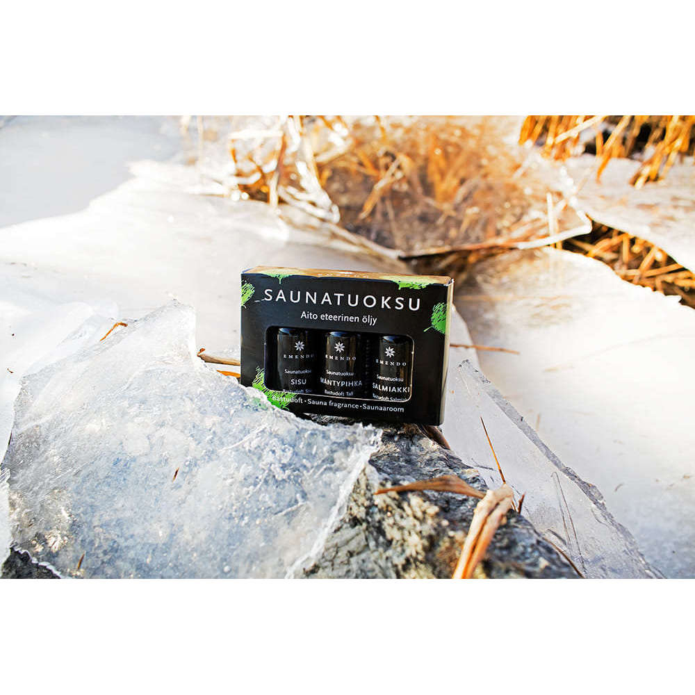 Una confezione di Emendo Sauna Fragrance Salmiac, Pine & Sisu di Emendo, prodotta in Finlandia, riposa all'aperto su ghiaccio crepato tra canne secche con la luce del sole che splende alle spalle.