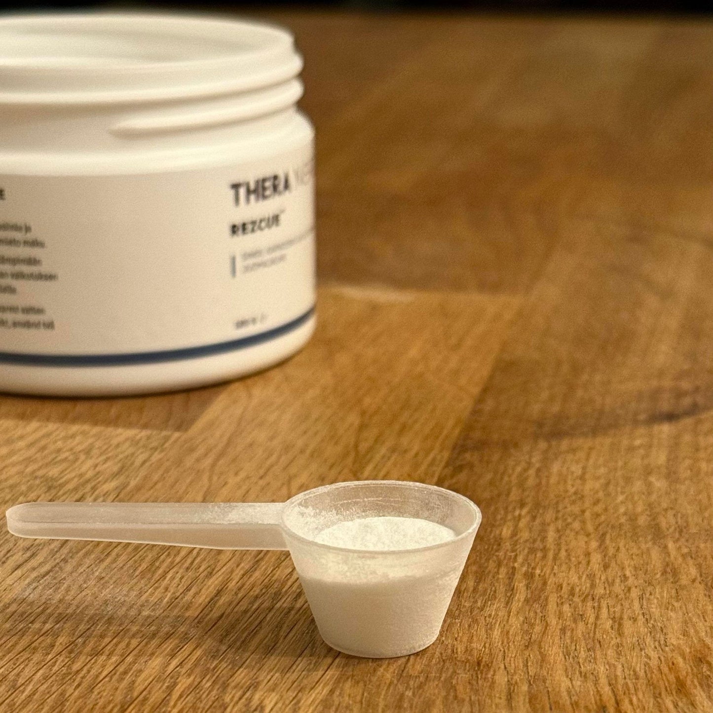 Een kleine plastic schep gevuld met wit THERA Nordic Rezcue Drink Powder staat op een houten oppervlak, wat verwijst naar de heilzame werking voor de darmen, met op de achtergrond een gedeeltelijk zichtbare THERA Nordic-verpakking.