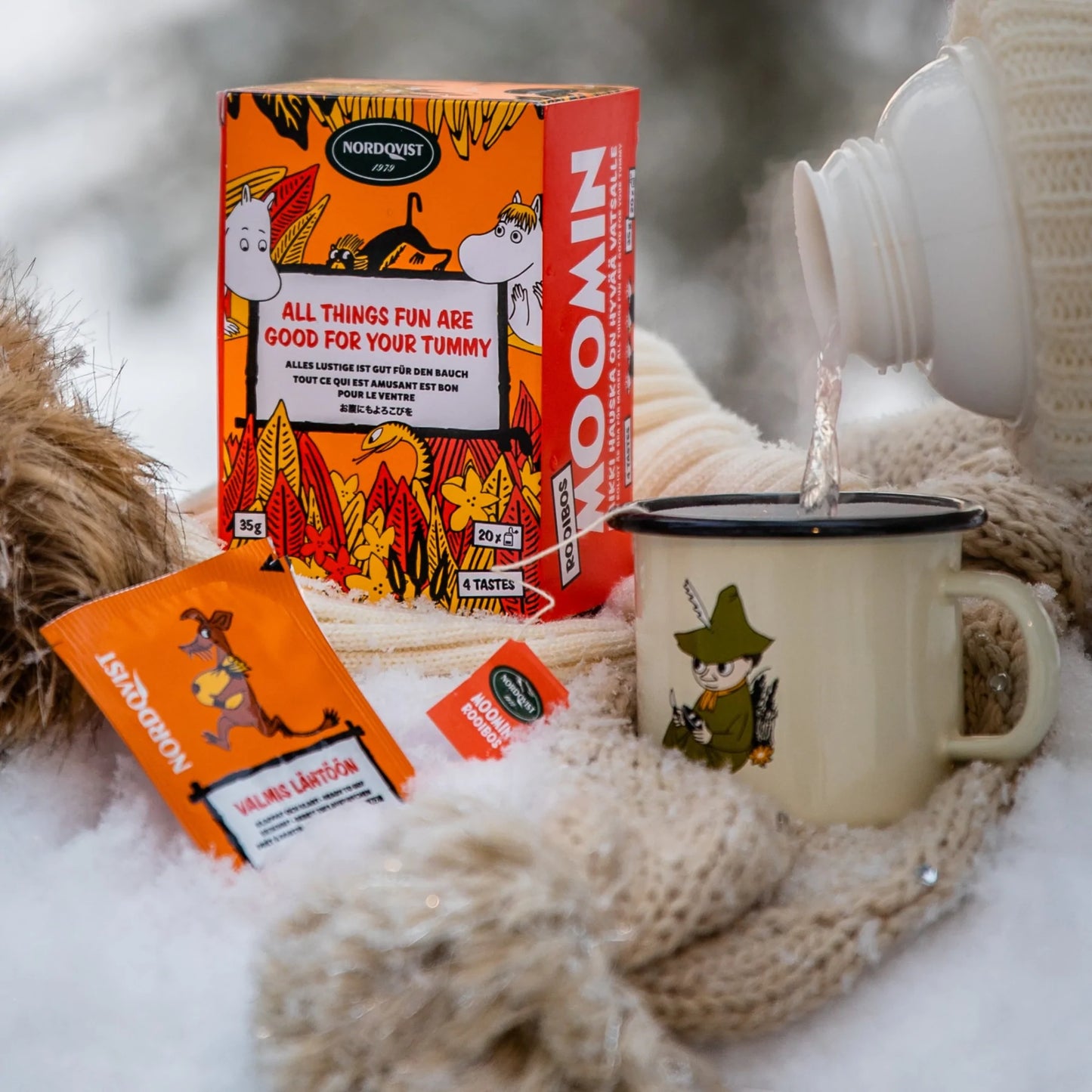 Gehuld in warme winterkleding schenkt iemand een warm drankje uit een thermoskan in een Snufkin-mok naast een doos en pakjes Nordqvist All Things Fun Are Good For Your Tummy rooibosthee, allemaal neergezet op de sneeuw.