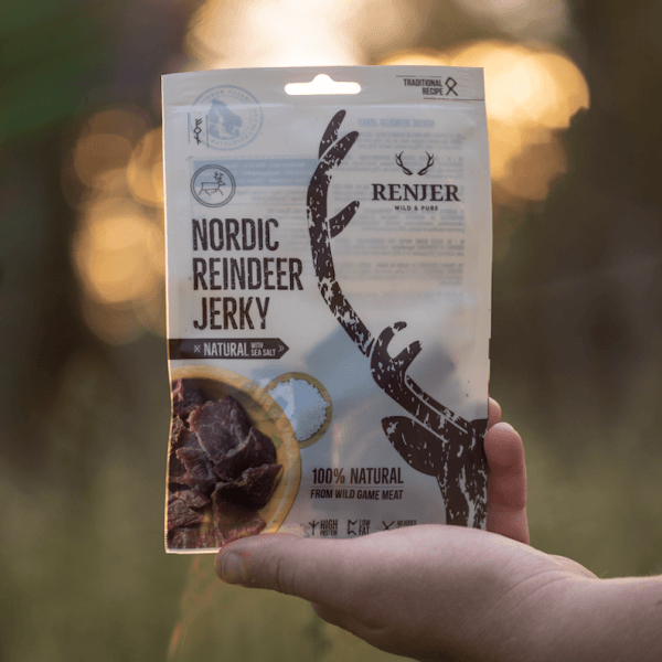 Een hand houdt een Renjer Nordic Reindeer Jerky Sea Salt-pakket vast in de buitenlucht, met daarop een rendiergewei, de tekst '100% natuurlijk' en een afbeelding van gesneden rendiervlees in een kom.