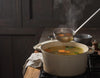 Golden bone broth being ladled from a steaming pot with herbs and vegetables