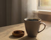 A steaming cup of coffee beside a small dish of dark mushroom coffee powder on a wooden desk