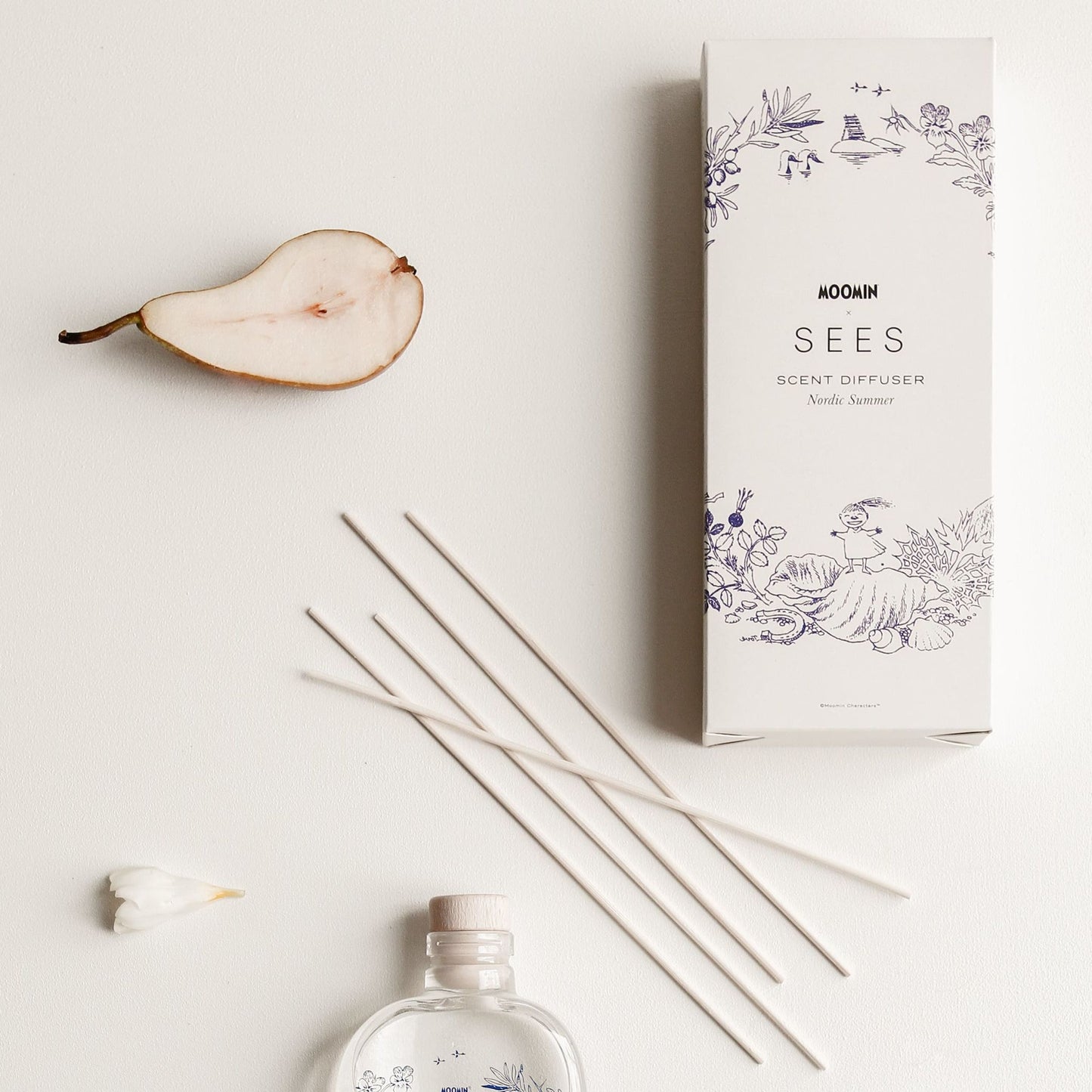 A flat lay of the SEES x Moomin Room Diffuser Nordic Summer by SEES Company, showing a decorative box, labeled glass bottle, four diffuser sticks, half a pear, and white flower petals on a white background.