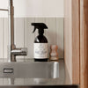 A close-up of a kitchen sink with a silver faucet, SEES Company’s SEES x Moomin Dish Wash Spray featuring a vegan formula, and a small wooden dish brush on the countertop against a tiled backsplash.