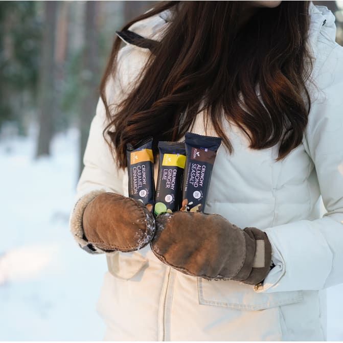 A person in a white winter jacket and brown mittens stands in a snowy forest, holding three Foodin Collagen Protein Bar Crunchy Almond Seasalt bars against their chest. Only the lower part of their face and hair are visible.