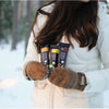 A woman in a white winter coat and brown mittens stands in a snowy forest, holding three Foodin Collagen Protein Bar Crunchy Cashew-Vanilla bars. Her face is not visible.