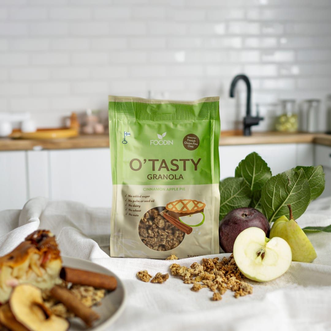 A bag of Foodin O`Tasty Granola Cinnamon Apple Pie stands on a white cloth, surrounded by granola pieces, apple slices, a whole apple, cinnamon sticks, and leaves—all made with gluten-free Finnish oats.