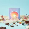 A square chocolate gift box labeled 'Natural Focus' stands on a light blue surface with scattered chocolate pieces, cream fabric, small flowers, and a watercolor palette. The box has a pastel gradient cover with a circular sun design and text reading 'Goodie Chocolate Company' and 'Vegan Chocolate'.