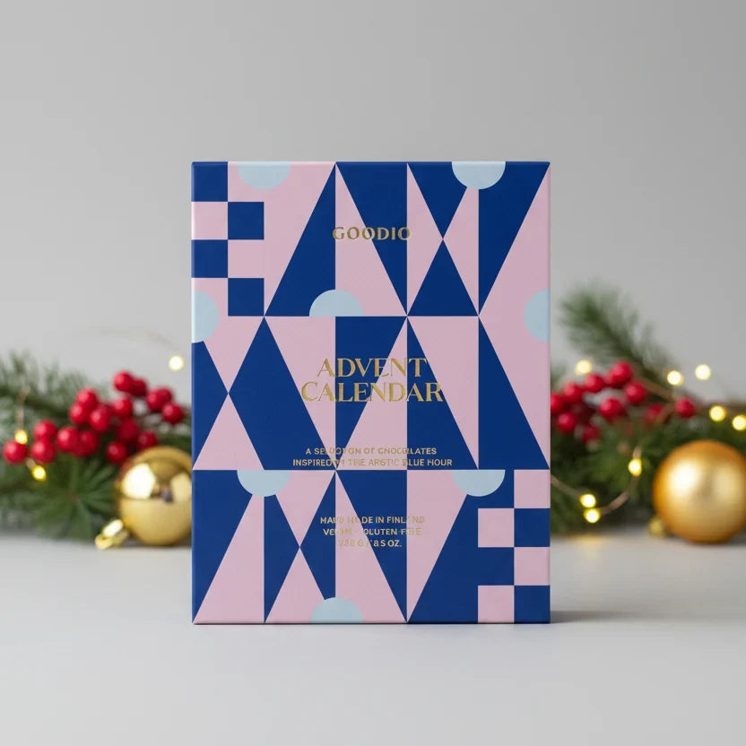 A Goodio Advent Calendar box with a geometric pattern of navy blue, pink, and light blue triangles, standing upright on a light gray surface with blurred Christmas decorations in the background.