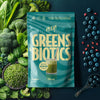 Package of 'HEEY! Greens & Biotics' surrounded by green vegetables and berries on a dark background