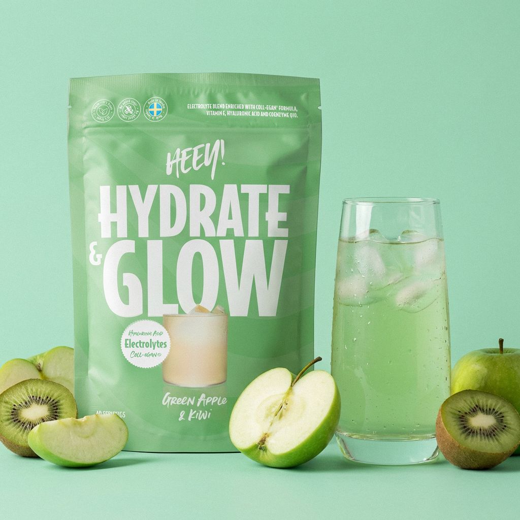 A green stand-up pouch of HEEN! Hydrate & Glow electrolyte drink mix stands next to a tall clear glass filled with a pale green iced drink, with sliced kiwi fruit arranged around them on a mint green background.