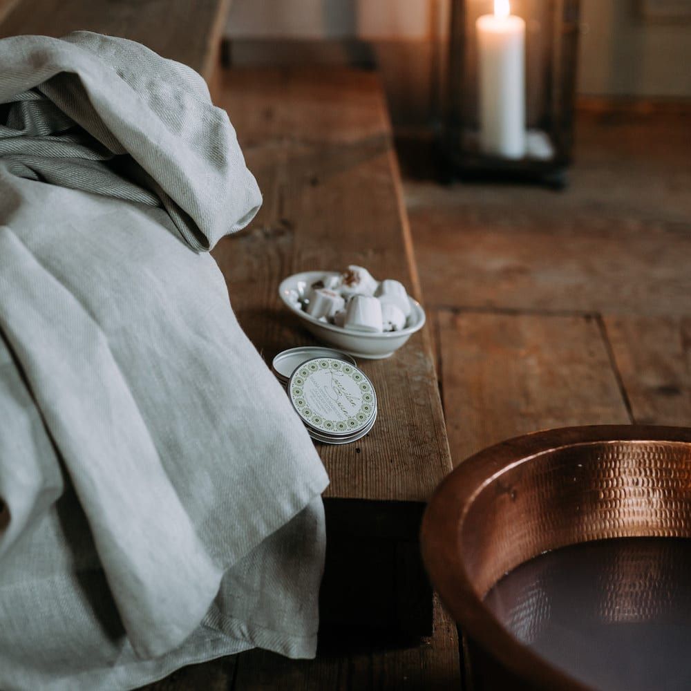A cozy spa scene with a folded linen towel, Kaurilan Sauna Grapefruit Footbath, and soaps on a wooden bench. A lit candle in a lantern glows beside a copper basin on the rustic wooden floor.