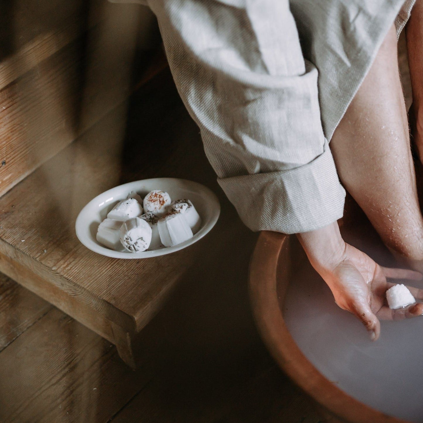 A person in a robe soaks their feet in milky water using the Kaurilan Sauna Peppermint Footbath, holding a white bath bomb. More Kaurilan Sauna vegan product bath bombs are placed on a plate on the wooden floor nearby.