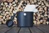 The Emendo Kolo Sauna Bucket & Ladle, featuring a black bucket with a metal handle and matching ladle, sits on wood alongside a white towel and stacked firewood—essential Nordic-inspired sauna accessories.