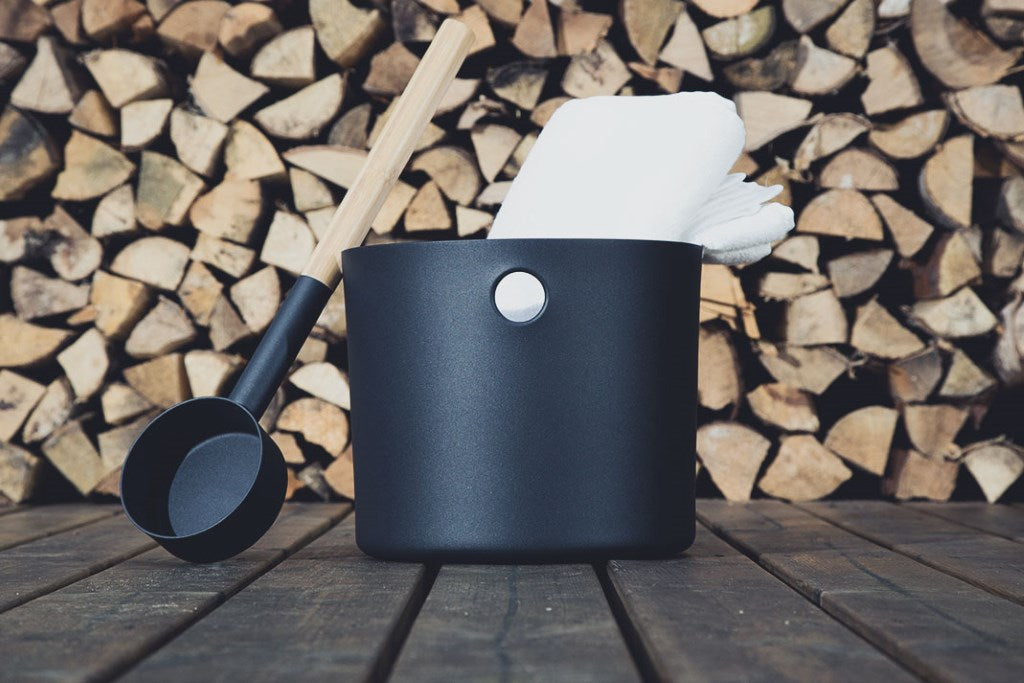 The Emendo Kolo Sauna Bucket & Ladle, featuring a black bucket with a metal handle and matching ladle, sits on wood alongside a white towel and stacked firewood—essential Nordic-inspired sauna accessories.