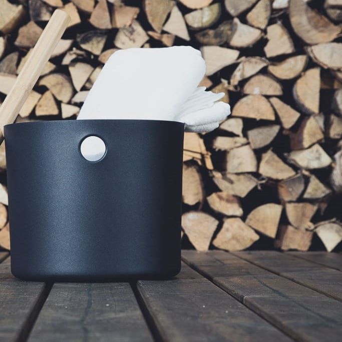 The Emendo Kolo Sauna Bucket & Ladle, featuring a black bucket with a metal handle and matching ladle, sits on wood alongside a white towel and stacked firewood—essential Nordic-inspired sauna accessories.