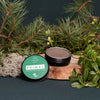 An open Luonkos Primal Multifunctional Forest Balm sits on a wooden slab among pine branches, green leaves, and moss—spotlighting its natural ingredients by Luonkos for eczema-prone skin—set against a dark backdrop.