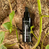 A black bottle of MoiForest Forest Glow Serum placed on textured tree bark with green leaves and vines around it.