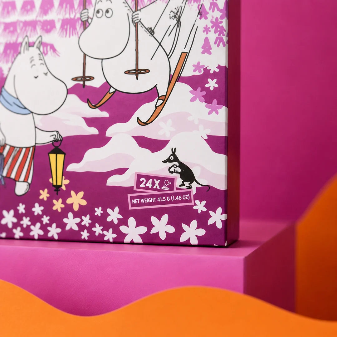 A close-up of a Moomin-themed product box in bright magenta packaging with illustrated characters, flowers, and snowy motifs. The box includes visible text reading '24 X' and 'NET WEIGHT 415 G (1.