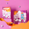 Two Moomin Tea Calendar boxes displayed on a pink studio background with orange paper wave shapes and floating tea bags. A glass of amber tea and a tea bag are placed in the foreground.