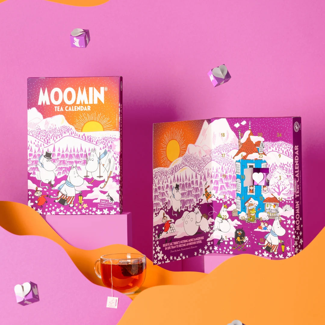 Two Moomin Tea Calendar boxes displayed on a pink studio background with orange paper wave shapes and floating tea bags. A glass of amber tea and a tea bag are placed in the foreground.