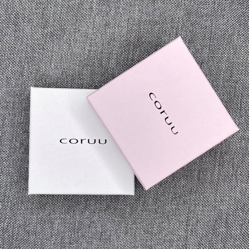 Two square boxes—one white and one light pink—both labeled "Coruu" in black, rest on gray fabric. The pink box slightly overlaps the white, displaying stylish packaging for Coruu Moomin Wooden Earrings.