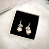 Coruu Moomin Wooden Earrings by Coruu, shaped like smiling cartoon characters in dresses, are displayed in a white jewelry box with black velvet interior. Nearby, a partially visible book titled Five Minute Journal can be seen.