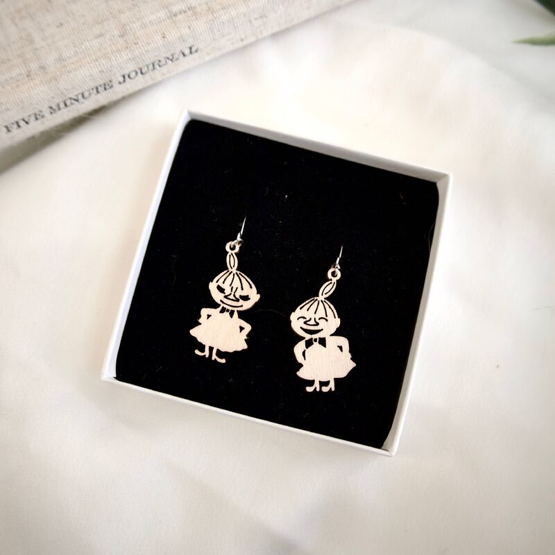 Coruu Moomin Wooden Earrings by Coruu, shaped like smiling cartoon characters in dresses, are displayed in a white jewelry box with black velvet interior. Nearby, a partially visible book titled Five Minute Journal can be seen.