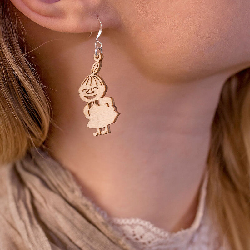 A close-up of a person with light skin and blonde hair wearing Coruu Moomin Wooden Earrings—a unique Nordic jewelry piece shaped like a smiling cartoon girl with a bun, hands behind her back.