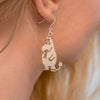 A close-up shows a person's ear wearing Coruu Moomin Wooden Earrings, shaped like the beloved cartoon character with a round body and small flowers, crafted from Finnish birch by Coruu, set against a blurred background.