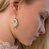 Close-up of a woman with light hair looking down, wearing Coruu Moomin Wooden Earrings shaped like a cartoonish monster from Coruu. The playful earrings are crafted from wood and feature a mischievous Moomin character.