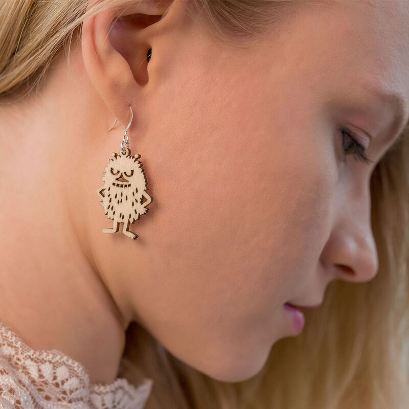 Close-up of a woman with light hair looking down, wearing Coruu Moomin Wooden Earrings shaped like a cartoonish monster from Coruu. The playful earrings are crafted from wood and feature a mischievous Moomin character.
